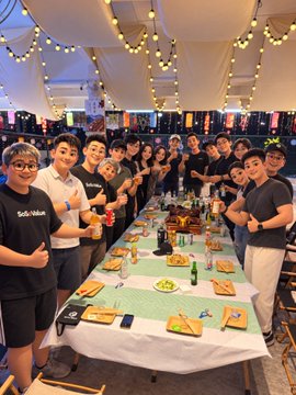 A lively group of attendees gathers around a long dining table during a SoSoValue-sponsored meetup at TOKEN2049 Singapore, with several wearing black "SoSoValue" t-shirts. Festive string lights and decorations set a social, community-focused atmosphere as smiling participants enjoy the evening together.