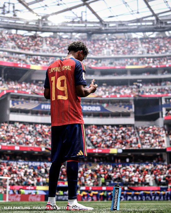 Photograph of soccer player Lamine Yamal (#19) in a red Spain jersey standing on the pitch during a match, with a Powerade bottle beside him and a full stadium in the background.