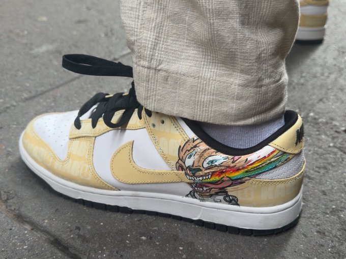 A pair of gold and white Nike Dunk Low sneakers featuring a cartoon character with a rainbow tongue graphic, paired with beige textured pants.