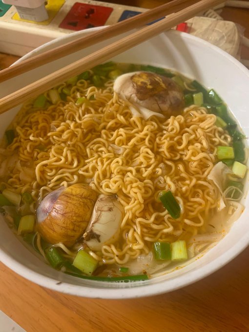 A close-up photo of a bowl of instant ramen noodles in broth with green onions and two whole boiled eggs, with wooden chopsticks resting on top.