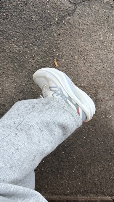 A first-person view looking down at someone's leg wearing gray sweatpants and a white, chunky-soled sneaker with red accents, standing on cracked asphalt.