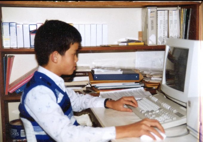 A young boy sits at an older computer, his hands on a white keyboard, surrounded by shelves full of binders and books. The image humorously suggests a very early glimpse of Binance founder CZ, playfully paired with the caption about him planning his future apartment sale to fund Binance.