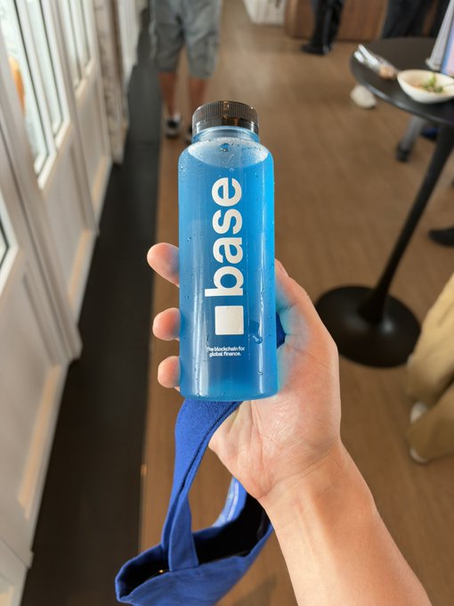 A hand holds a clear bottle filled with bright blue liquid, branded "base" with the tagline "The blockchain for global finance" printed on the label, visually representing the "base juice" mentioned in the post as an energizing promotional drink for the Base blockchain.