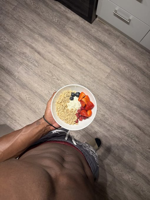A shirtless person holds a bowl of granola, blueberries, yogurt, and strawberries in a kitchen, likely showcasing a healthy post-workout meal as a nighttime routine nod to the fitness-focused "Gnight fam" message.