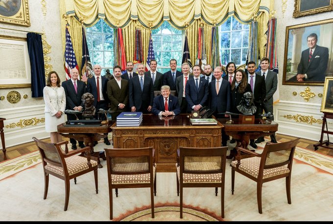 President Trump is seated at the Resolute Desk in the Oval Office, surrounded by a group of officials and advisors posing for a formal group photo. The accompanying post frames the image as a cryptic signal that the administration is preparing to integrate XRP and Ripple into the new U.S. financial system.
