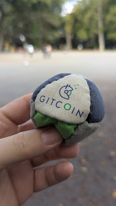 A hand holds a small, round plush toy designed to look like a Gitcoin logo button on an outdoor street.