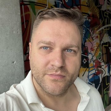 A selfie of a man with short hair and light eyes wearing a white collared shirt, standing in front of colorful street art.