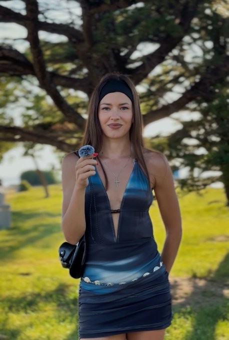 The image features a woman wearing a blue dress with a plunging neckline, standing outdoors under a tree. She holds an ice cream cone in her hand while posing for the picture. The background of the photo includes trees, suggesting that they are in a park or a similar outdoor setting. The caption provided seems to be unrelated and does not provide any context about the image or its content.