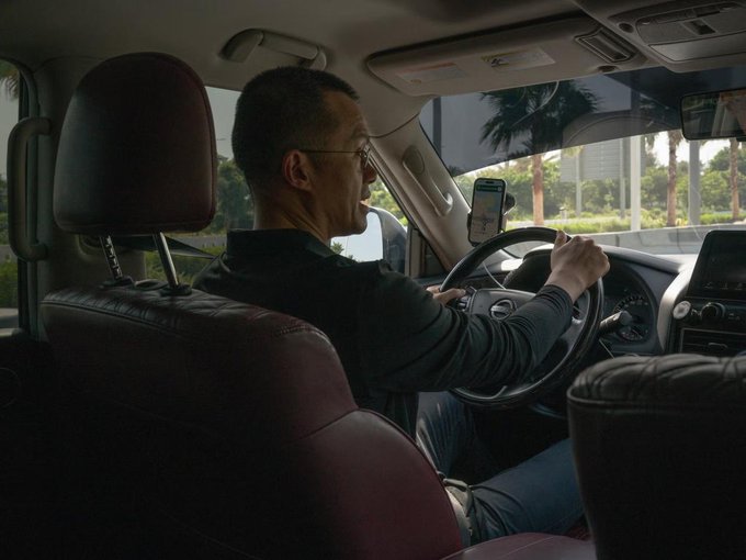 A man sits behind the wheel of a car with a phone mounted on the dashboard displaying a navigation app, appearing to work as a rideshare driver. The image satirically illustrates the post's claim by visually depicting the "former kingpin" humbly disguised as an everyday Uber driver seeking sympathy.