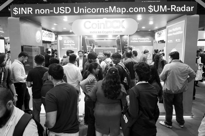 A black and white image showing a crowded crypto conference booth for CoinDCX with text 'India ka Crypto Coach' and '0.01% Trading Fee', overlaid with text reading '$INR non-USD UnicornsMap.com $UM-Radar'.