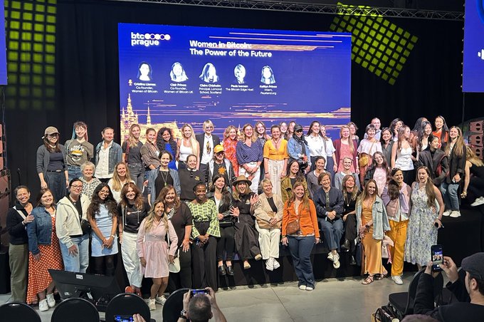 The image shows a large group of people posing together, likely at an event or gathering. The group consists of both men and women, all dressed similarly with colorful clothing. They are standing close to each other, creating a sense of unity and camaraderie among them. The background features a stage setup with a screen displaying the words "Women in Business" written on it. This suggests that this event is focused on celebrating and promoting women's achievements in business or professional settings.
