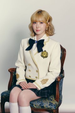 A blonde-haired woman sits in an ornate chair wearing a cream-colored school uniform jacket with gold embroidery and a navy bow tie.
