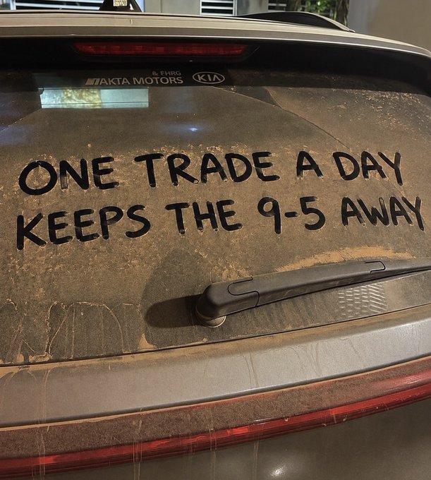A dusty car rear window with 'ONE TRADE A DAY KEEPS THE 9-5 AWAY' written in the dirt, with 'AKTA MOTORS' and 'KIA' dealership decals visible.