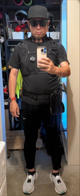 A man wearing a black cap, sunglasses, tactical vest, and black athletic wear takes a mirror selfie in front of a display case of helmets.