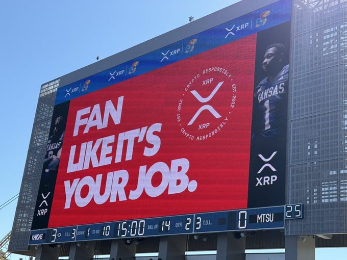 A stadium scoreboard displaying a red XRP cryptocurrency advertisement with the text 'FAN LIKE IT'S YOUR JOB.' along with sports scores.