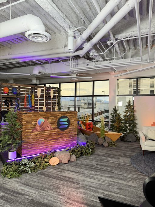 An office reception area decorated with a rustic wooden desk, potted plants, artificial trees, rocks, and a canoe, set under exposed industrial ceiling pipes.