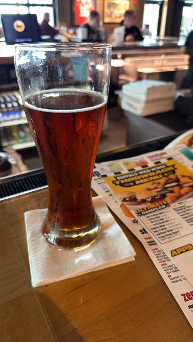 A tall pilsner glass filled with amber beer sits on a napkin atop a wooden bar counter, next to a Buffalo Wild Wings menu displaying "WINGS THAT BRING THE HEAT" and a list of apps.