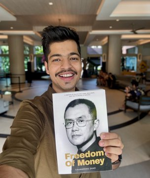 A selfie of a man holding up a signed copy of Changpeng Zhao's "Freedom of Money" book at Binance Campus Asia, with visible text on the cover confirming the charity aspect of the book royalties.