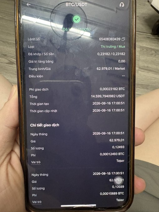 A hand holds a smartphone displaying a Vietnamese-language trading app showing a completed BTC/USDT buy order for 0.12493 BTC at a price of 62,979.01, dated 2026-08-16.