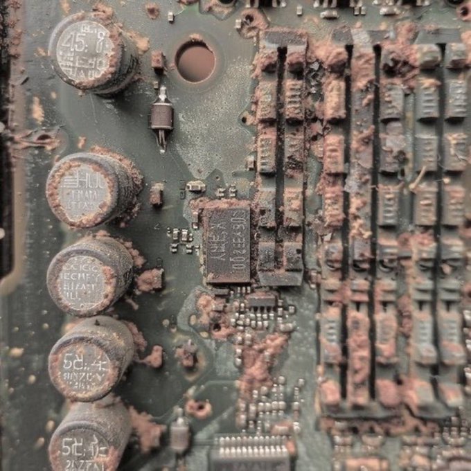 A close-up photograph of a dusty circuit board featuring cylindrical capacitors and an AMD chip, heavily coated in a reddish-brown residue.