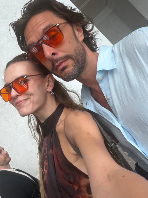 A close-up selfie of a man and woman both wearing matching orange-tinted sunglasses, with the man in a light blue shirt and the woman in a sheer brown top.