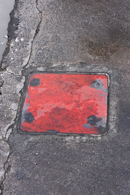 A worn, weathered red square metal cover embedded in cracked asphalt pavement.