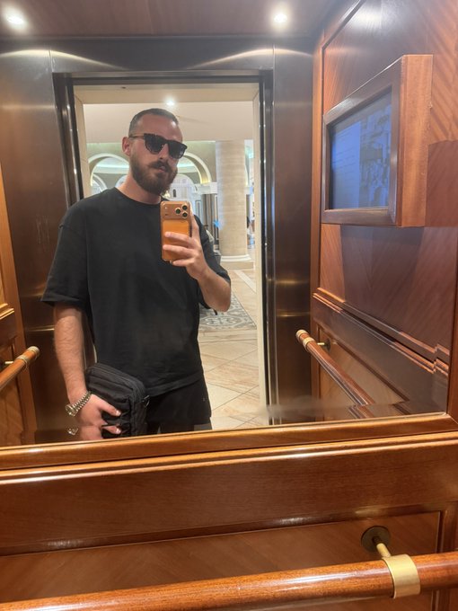 A stylish man in sunglasses and a black t-shirt takes a mirror selfie with his phone inside a wood-paneled elevator, with a small screen mounted on the wall beside him. The "vibe check" caption perfectly matches his effortlessly cool, confident demeanor as he casually documents the polished, upscale atmosphere of the ride.