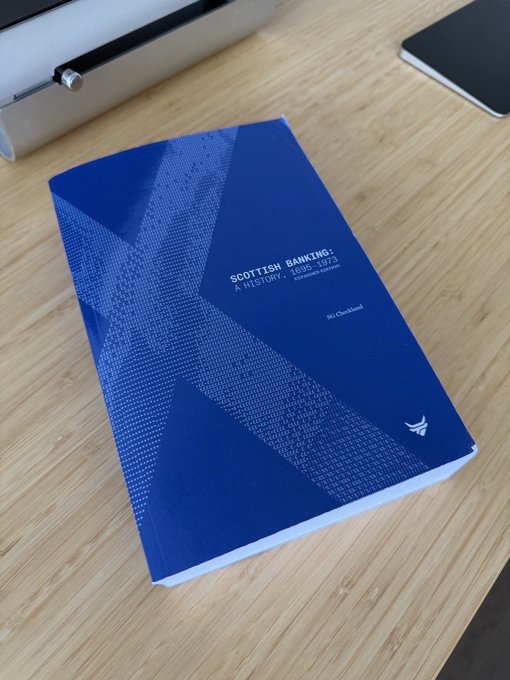 A blue book titled "Scottish Banking: A History, 1695-1973" by S. G. Checkland rests on a wooden desk, its substantial thickness visible from the side, making it a dense and intriguing read about the history of banking in Scotland.