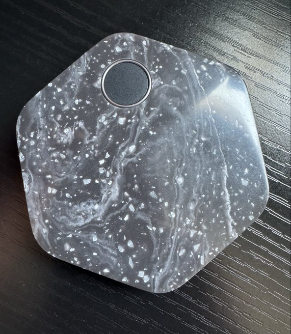 A hexagonal resin object with a swirling black and white marbled pattern and speckled details, featuring a circular black center, rests on a dark wood surface.