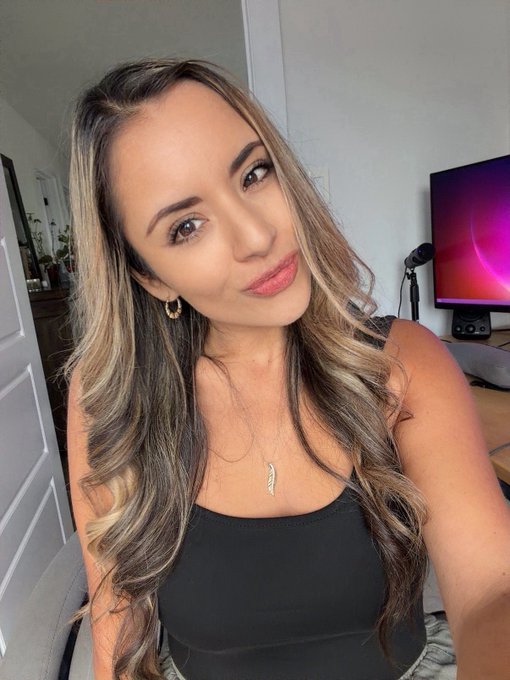 A woman with long blonde-highlighted hair wearing a black tank top and a feather pendant necklace poses for a selfie, with a studio light and computer monitor visible in the background.