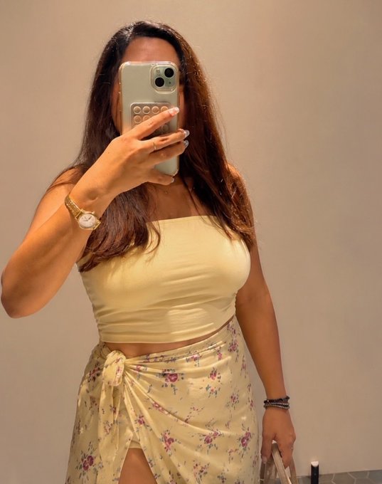 A woman in a pale yellow strapless top and matching floral wrap skirt takes a mirror selfie with her phone, her face obscured by the device.