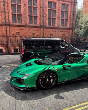 A bright green Ferrari sports car parked on a city street in front of a red brick building with a black van parked behind it.