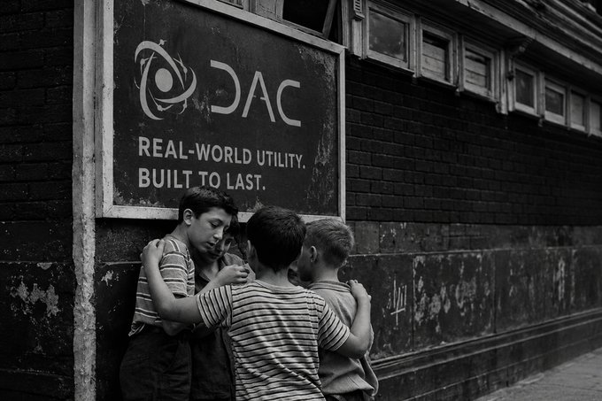 A black and white photograph of four young boys huddled together against a brick wall beneath a sign reading 'DAC REAL-WORLD UTILITY. BUILT TO LAST.'