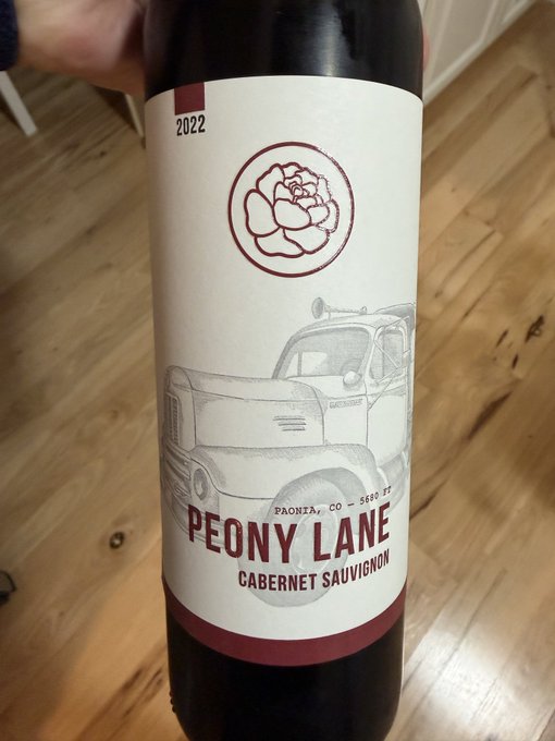 A bottle of 2022 Peony Lane Cabernet Sauvignon from Paonia, CO, featuring a white label with a peony flower emblem and illustration of a vintage truck, held against a wooden floor background.