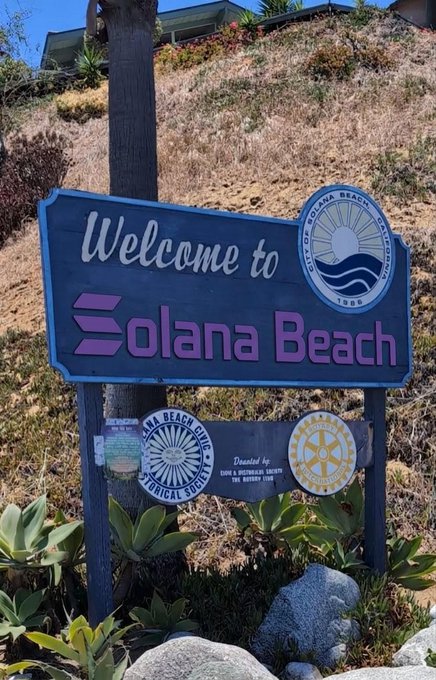 The image features a blue sign that reads "Welcome To Solana Beach" with a picture of a beach, surrounded by plants and rocks. There are also two stickers on the sign, which seem to be related to the Crypto movement or blockchain technology. The text in the image suggests that the name Solana originally referred to a specific beach, but over time it has become associated with a broader concept or movement within the crypto world.