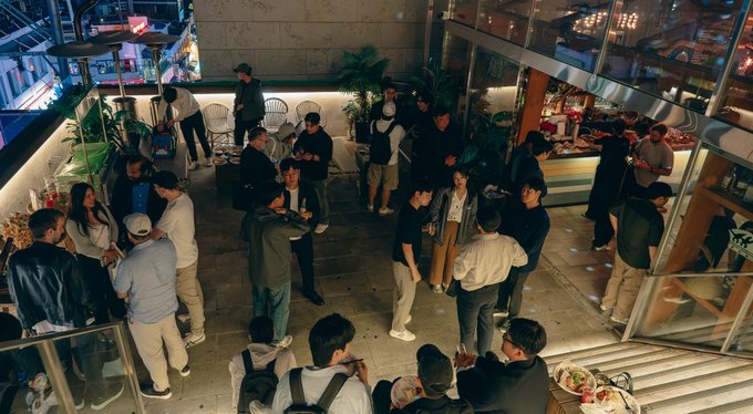 A nighttime photo showing a large gathering of people mingling in what appears to be an outdoor venue or patio area, with guests holding drinks and conversing in small groups.