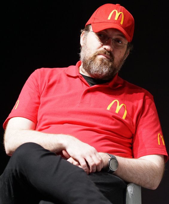 The image features a man wearing a red shirt with a McDonald's logo sitting down. He has glasses on his face and appears to be deep in thought or contemplation, as he looks off into space. The photo does not provide any information about the content of the image being related to Charles Hoskinson's statement about decentralization.