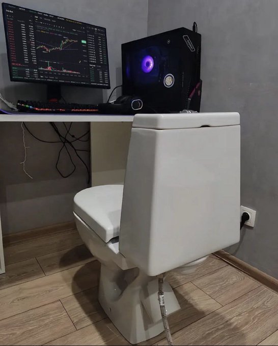 A photograph of an unusual bathroom setup featuring a white toilet positioned in front of a desk with a computer monitor displaying trading charts, a backlit PC tower with purple lighting, and a gaming keyboard.