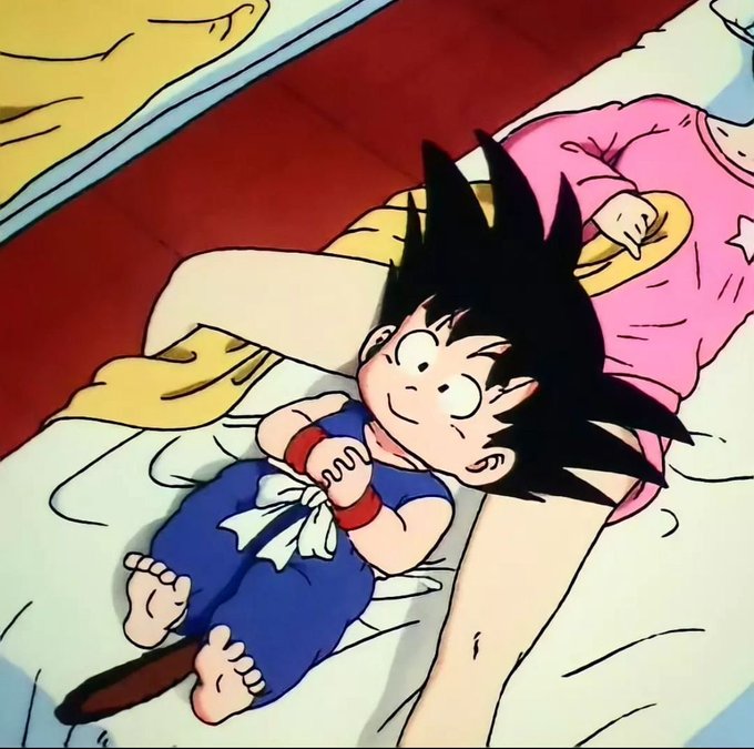 An anime illustration of young Goku lying in bed next to his mother, wearing a blue outfit with a white sash and red wristbands, with his tail visible.