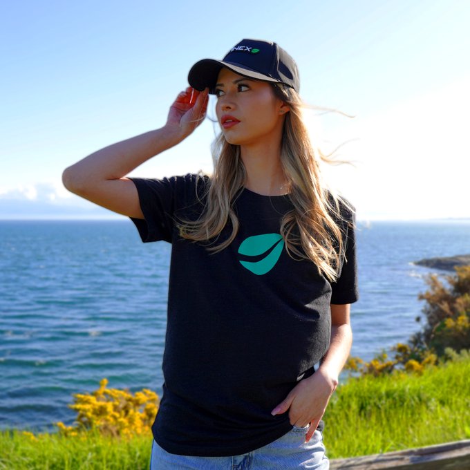 A young woman with long blonde hair wearing a black cap and a black t-shirt, both branded with the Teneo logo, posing outdoors against a coastal backdrop with blue ocean and sky.