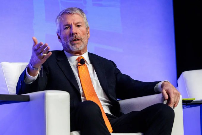 An older man with gray hair and a beard, wearing a black suit and orange tie, speaking while gesturing with his hand while seated in a white chair on stage.