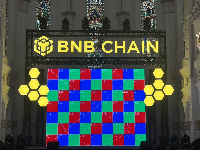 An event display showing the 'BNB CHAIN' logo above a colorful numbered grid and hexagonal decorations inside what appears to be a venue with stained glass windows.