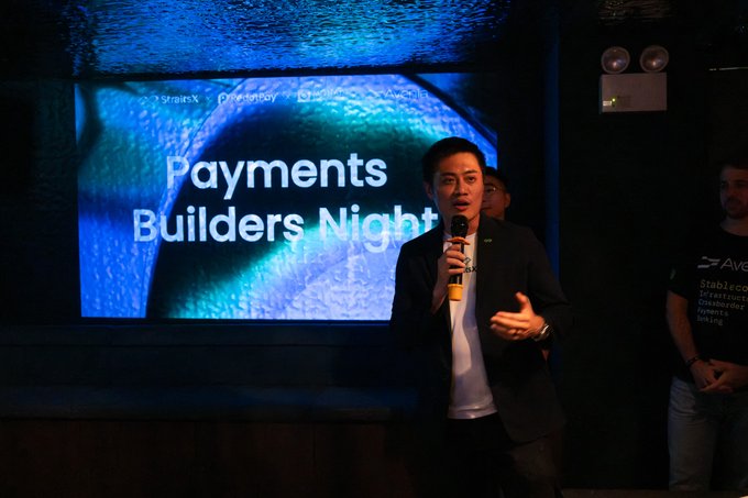 A man in a dark blazer speaks into a mic on stage at 'Payments Builders Night' with a large blue screen behind him displaying the event title.