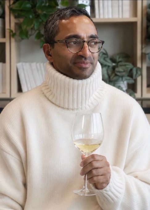 A man in a cream turtleneck sweater and glasses holds a glass of white wine in front of a bookshelf.