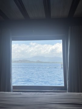 View from inside a bedroom showing a large window overlooking an ocean and distant mountains, with white curtains framing the scene and a bed in the foreground. The image complements the "GM my x frens i woke up like this" message, illustrating a peaceful, scenic morning wake-up view.