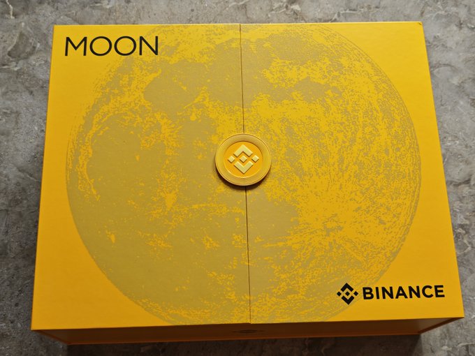 A yellow Binance-branded box labeled 'MOON' featuring a Binance logo seal in the center, set against a marble background.