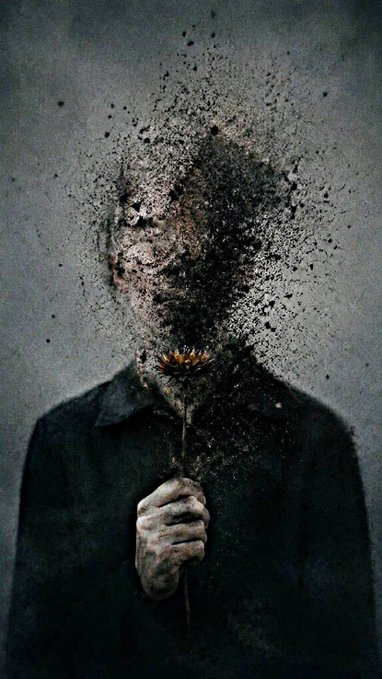 A dark, surreal artistic image of a person whose head and upper body appear to be disintegrating into a cloud of black particles and dust, holding what looks like a dried flower.