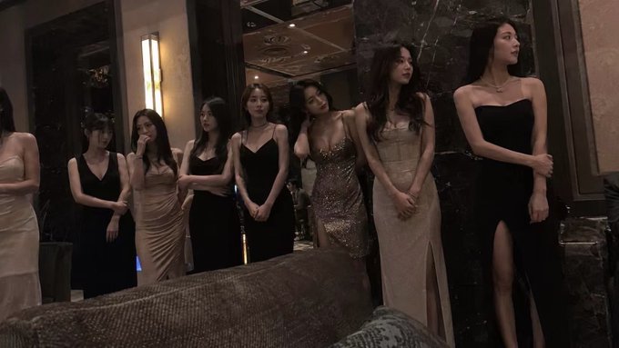The image features a group of six women standing together, all dressed up for an event or party. They are wearing long dresses that cover their entire bodies except for their hands and feet. The room they are in appears to be a dark space with marble walls, giving off an elegant atmosphere. One woman is leaning against the wall while the others stand close by, possibly engaged in conversation or waiting for something.