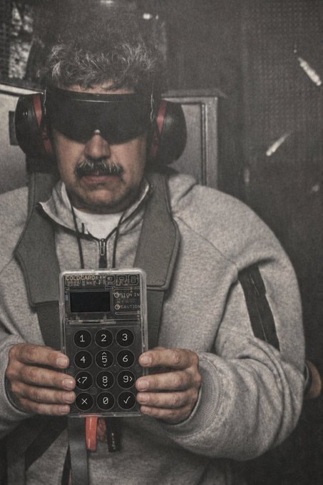 A man wearing dark goggles and red-and-black earmuffs holds a vintage battery-testing meter labeled 'CONDUCTRONICS BRT-10' in his gloved hands.