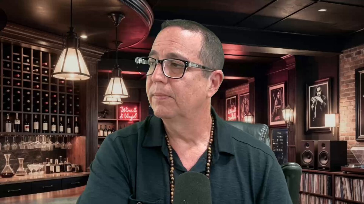 The image features a man wearing glasses sitting at a bar, with several wine bottles behind him. The background includes a dark room with a brick wall and a ceiling lamp hanging from the ceiling. There are also some pictures hanging on the wall above the man's head. The text "25% Of G20 Nations On XRP youtu.be/SQCelArIEUY" is present in the image, which could be related to the post or just part of the scene.